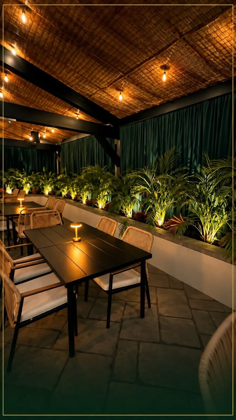 A private table with a brass lamp, palms lit from below, and emerald curtains